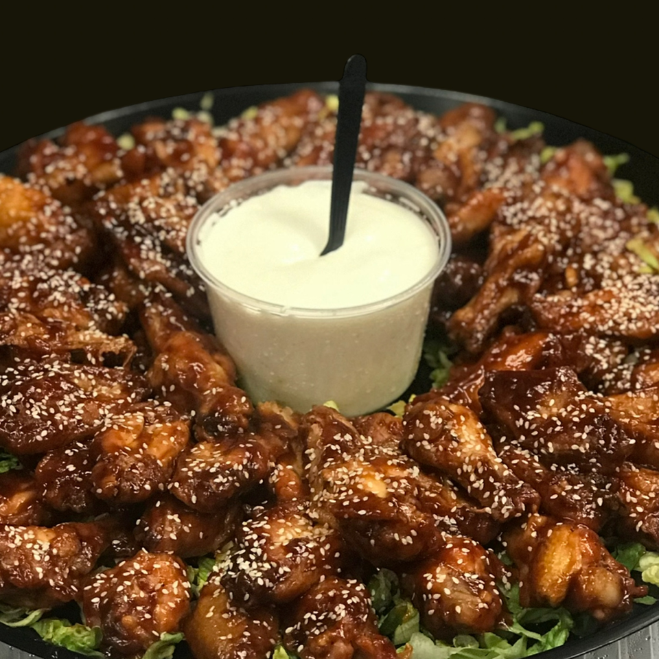 A platter of sesame chicken wings with a side of creamy white dipping sauce and lettuce.