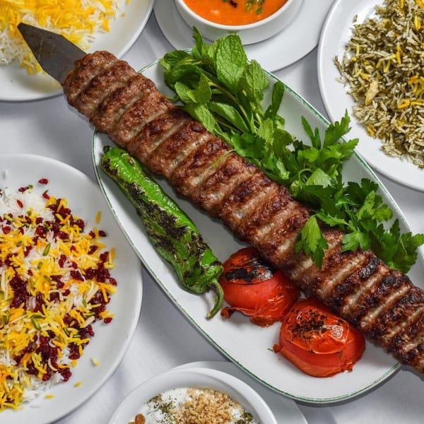A long Persian kabob with grilled vegetables and herbs, surrounded by rice dishes and soup.