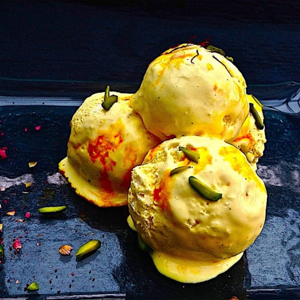 Three scoops of saffron ice cream, garnished with pistachios and red streaks, on a dark surface.