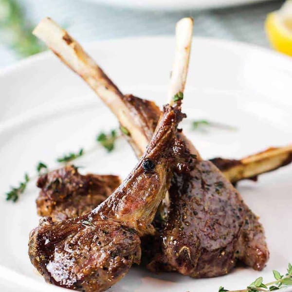 Three succulent lamb chops, seasoned and grilled, garnished with fresh thyme on a white plate.