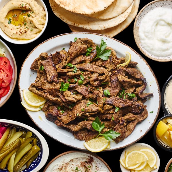 Platter of grilled meat with lemon slices, surrounded by hummus, pita, yogurt, pickles, and tomatoes.