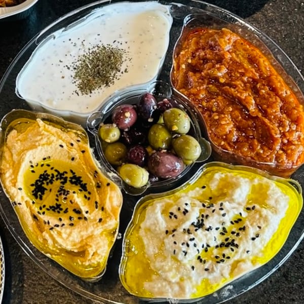 A clear platter with four dips: white yogurt with herbs, red relish, hummus, and white dip with oil and seeds, plus olives.