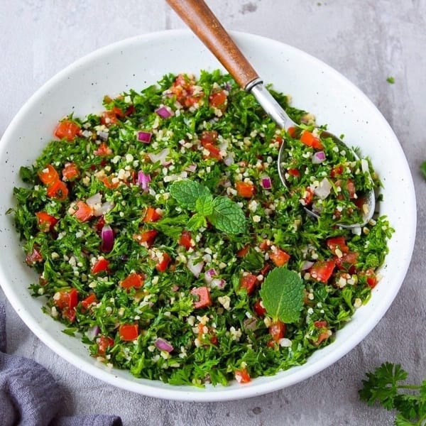 A white bowl filled with vibrant green tabbouleh salad, garnished with mint, and a serving spoon.