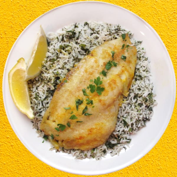 Golden-brown fish fillet with parsley, served on a bed of herb rice with lemon wedges.